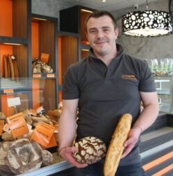Adrien, boulanger et gérant d'un distributeur automatique, pose dans son magasin avec du pain dans les mains